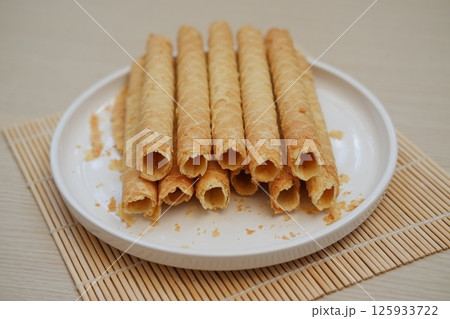 The image shows a plate of crispy, golden-brown wafer rolls neatly arranged in two layers on a white ceramic plate The image shows a plate of crispy, golden-brown wafer rolls neatly arranged in two layers on a white ceramic plate 125933722