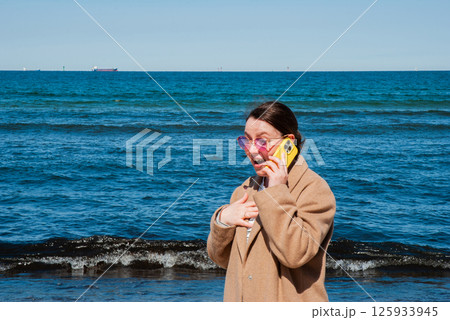 Young caucasian female talking on a yellow phone at the beach, wearing pink sunglasses and a brown coat, with the ocean in the background. Digital nomad lifestyle. Remote work 125933945