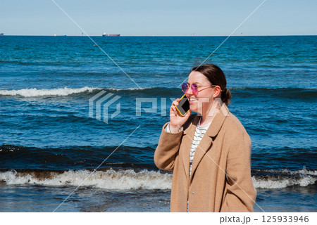 Young caucasian woman in a brown coat talking on a smartphone at the beach with blue ocean waves in the background on a sunny day. Digital nomad lifestyle. Remote work 125933946