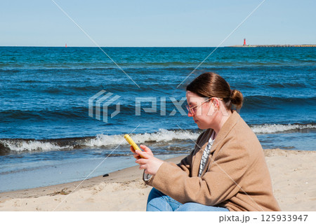 Young caucasian woman in casual attire enjoying a sunny day at the beach while browsing her smartphone, with ocean waves in the background. Digital nomad lifestyle. Remote work 125933947