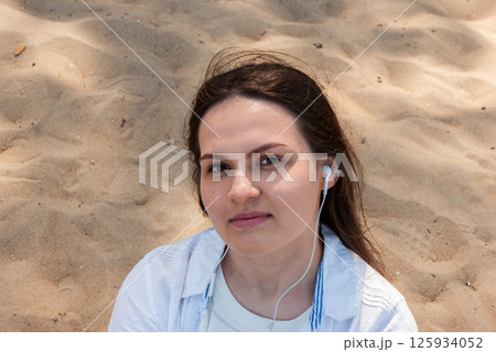 Young caucasian female with earphones at beachside, engaged in relaxing moment, wearing casual attire, surrounded by sandy shores. Digital nomad lifestyle. Remote work Young caucasian female with earphones at beachside, engaged in relaxing moment, wearing casual attire, surrounded by sandy shores. Digital nomad lifestyle. Remote work 125934052