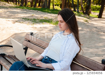 Young caucasian woman using laptop on a sunny park bench with earphones, multitasking outdoors on a serene sandy path amidst lush green trees. Digital nomad lifestyle. Remote work 125934056