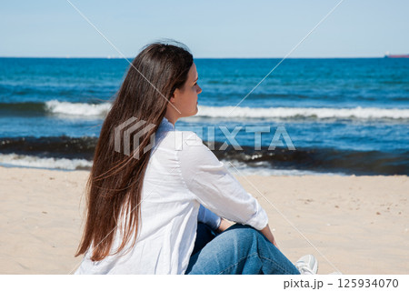 Young caucasian female with long brown hair in casual attire sitting on a sandy beach, gazing at the calm ocean waters under a clear blue sky. Digital nomad lifestyle. Remote work Young caucasian female with long brown hair in casual attire sitting on a sandy beach, gazing at the calm ocean waters under a clear blue sky. Digital nomad lifestyle. Remote work 125934070