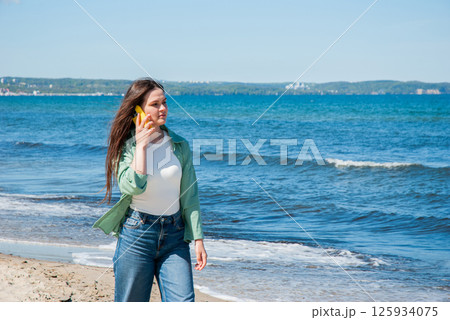 Young caucasian female walking by the ocean on a sunny day, talking on a smartphone, showcasing casual beach fashion and carefree moments. Digital nomad lifestyle. Remote work 125934075