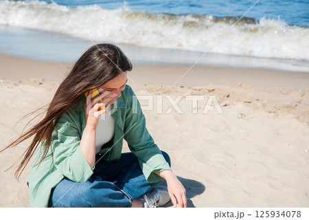 Young caucasian female enjoying a sunny day at the beach while talking on the phone, sitting on sand near ocean waves in casual green jacket. Digital nomad lifestyle. Remote work 125934078