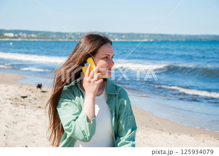 Young caucasian female enjoying a sunny beach day while talking on a smartphone with waves and blue sky in the background. Digital nomad lifestyle. Remote work 125934079