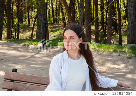 Young caucasian female relaxing on park bench in forested area during a bright day, wearing white shirt and looking thoughtful and serene. Digital nomad lifestyle. Remote work Young caucasian female relaxing on park bench in forested area during a bright day, wearing white shirt and looking thoughtful and serene. Digital nomad lifestyle. Remote work 125934084