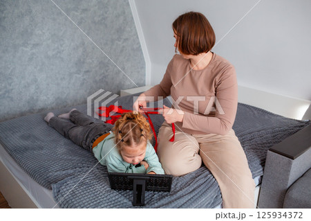 Top view of young mother braids her daughter's hair while sitting on the couch. A little girl uses a tablet 125934372