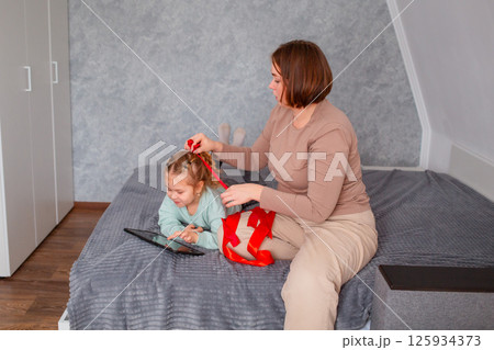 Wide shot of Caucasian woman braids the hair of a little girl. Mother and cute daughter relax on the bed and watch cartoons on tablet Wide shot of Caucasian woman braids the hair of a little girl. Mother and cute daughter relax on the bed and watch cartoons on tablet 125934373