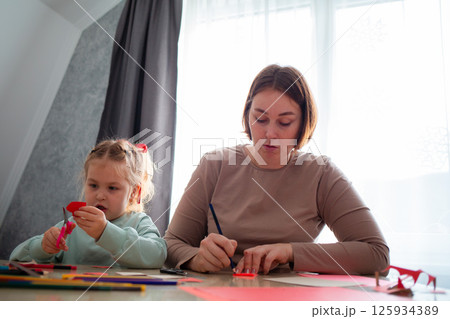 Low angle view of adult Caucasian mother and her little daughter cut out paper hearts sitting at a desk. Family is preparing for the celebration of Valentine's Day 125934389
