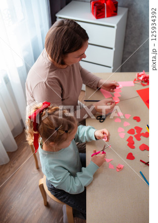 Top view of little cute girl and her mother cut out paper hearts sitting at a desk. Family is preparing for the celebration of Valentine's Day, Vertical 125934392