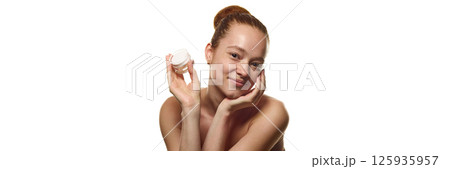 Portrait of beautiful young woman with spotless freckled skin holding jar of cream, touching cheek over white background. 125935957