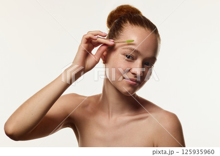 Portrait o young woman with natural skin and freckles, holding eyebrow brush near her eyebrow over white background. 125935960
