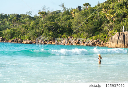 Woman in bikini and hat stands in turquoise sea at a tropical Seychelles beach with granite boulders Woman in bikini and hat stands in turquoise sea at a tropical Seychelles beach with granite boulders 125937062