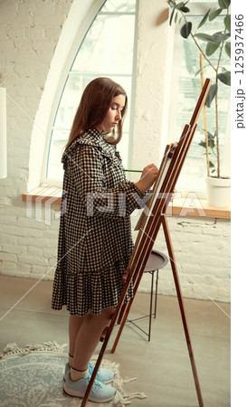 Teenage girl in dress standing at window and painting on easel with concentration on canvas Teenage girl in dress standing at window and painting on easel with concentration on canvas 125937466