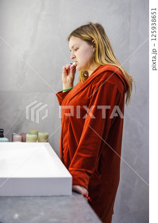 Young woman in a red bathrobe brushes her teeth while standing by the mirror, following a calm daily hygiene routine in a bright modern bathroom. Young woman in a red bathrobe brushes her teeth while standing by the mirror, following a calm daily hygiene routine in a bright modern bathroom. 125938651