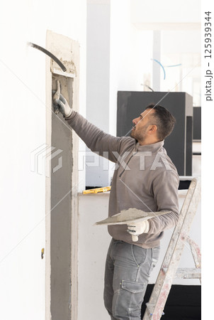 Craftsman in dirty gloves and gray clothes spreading plaster with a metal float over a doorway 125939344