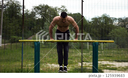 Hardy shirtless sportsman doing exercises on parallel bars at nature. Strong and muscular man working out at sports ground. Young athletic guy exercising outdoor. Concept of active healthy lifestyle Hardy shirtless sportsman doing exercises on parallel bars at nature. Strong and muscular man working out at sports ground. Young athletic guy exercising outdoor. Concept of active healthy lifestyle 125939683