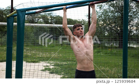 Young muscular sportsman doing pull ups at sports ground. Athletic guy exercising outdoor. Strong sporty man working out at summer nature. Concept of healthy and active lifestyle. Slow motion Young muscular sportsman doing pull ups at sports ground. Athletic guy exercising outdoor. Strong sporty man working out at summer nature. Concept of healthy and active lifestyle. Slow motion 125939684