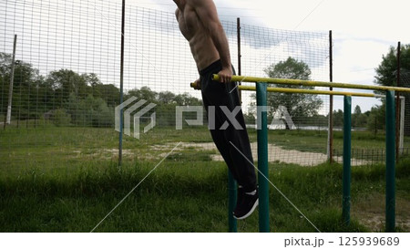 Young sportsman doing push ups on parallel bars at nature. Strong sporty man exercising at sports ground. Male shirtless athlete training hard outdoor. Concept of active sportive lifestyle. Slow mo Young sportsman doing push ups on parallel bars at nature. Strong sporty man exercising at sports ground. Male shirtless athlete training hard outdoor. Concept of active sportive lifestyle. Slow mo 125939689