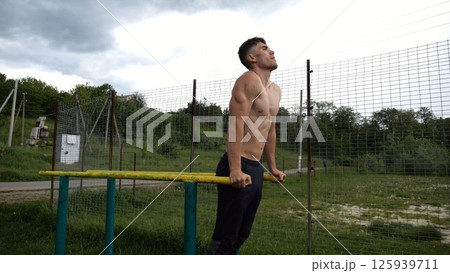 Young sportsman doing push ups on parallel bars at nature. Strong sporty man exercising at sports ground. Male shirtless athlete training hard outdoor. Concept of active sportive lifestyle. Slow mo Young sportsman doing push ups on parallel bars at nature. Strong sporty man exercising at sports ground. Male shirtless athlete training hard outdoor. Concept of active sportive lifestyle. Slow mo 125939711