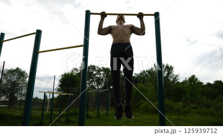 Muscular athlete doing pull ups on horizontal bar at sports ground. Young shirtless sportsman exercising at nature. Strong sporty man training outdoor. Concept of healthy and active lifestyle. Slow mo Muscular athlete doing pull ups on horizontal bar at sports ground. Young shirtless sportsman exercising at nature. Strong sporty man training outdoor. Concept of healthy and active lifestyle. Slow mo 125939712