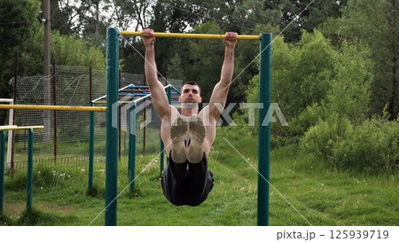 Athletic man trains abdominal muscles raising legs on horizontal bar at sports ground. Young sportsman exercising on pull-up bar outdoor. Sporty athlete working out at nature. Healthy lifestyle Athletic man trains abdominal muscles raising legs on horizontal bar at sports ground. Young sportsman exercising on pull-up bar outdoor. Sporty athlete working out at nature. Healthy lifestyle 125939719