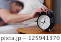 Exhausted man being awakened by an alarm clock in his bedroom 125941370