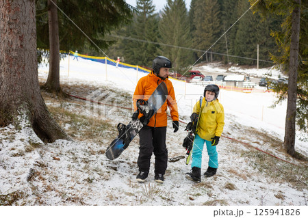 Winter adventures on a snow covered slope as a parent and child prepare for a day of snowboarding in a picturesque mountain landscape surrounded by tall trees Winter adventures on a snow covered slope as a parent and child prepare for a day of snowboarding in a picturesque mountain landscape surrounded by tall trees 125941826