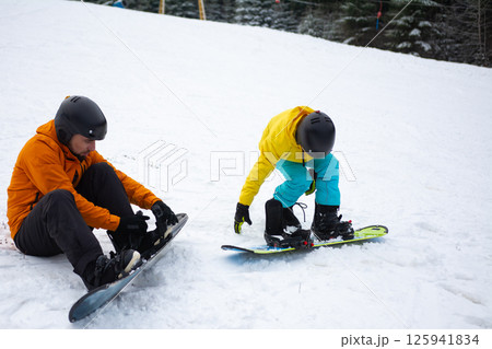 Adventurous snowboarders preparing for a thrilling descent on a snowy slope surrounded by beautiful evergreen trees during a bright winter day Adventurous snowboarders preparing for a thrilling descent on a snowy slope surrounded by beautiful evergreen trees during a bright winter day 125941834