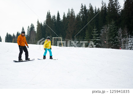 Winter adventure unfolds on a snowy slope as two snowboarders glide gracefully down the hill, surrounded by a tranquil forest of evergreen trees Winter adventure unfolds on a snowy slope as two snowboarders glide gracefully down the hill, surrounded by a tranquil forest of evergreen trees 125941835