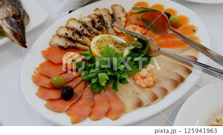 Gourmet catering platter displaying fresh seafood selection, including sliced fish, shrimp, garnished with lemon, parsley, olives on pristine white serving dish 125941963