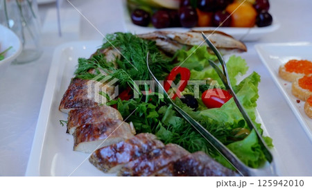 Grilled fish slices lying on white plate, accompanied by dill, lettuce, cherry tomatoes, black olives, bell peppers, presenting vibrant mediterranean style seafood arrangement 125942010