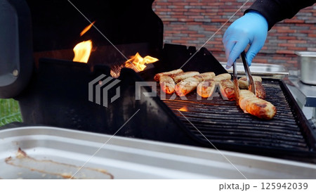 Chef wearing blue gloves uses tongs to grill sausages over flames on a barbecue grill during an outdoor party, ensuring they're perfectly cooked for guests 125942039