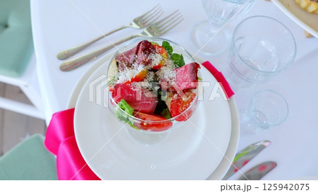 Hearty beef salad in glass bowl, layering crisp arugula, juicy tomatoes, creamy mozzarella, sharp parmesan over white plate with pink napkin, silverware nearby 125942075