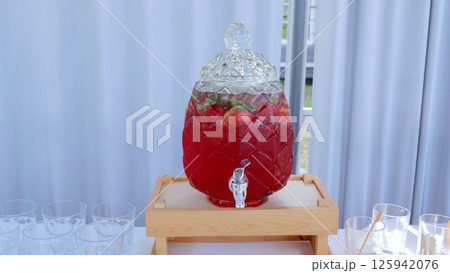 Glass beverage dispenser holding vibrant red fruit punch, garnishing with ice, mint, fresh fruit slices, resting on wooden stand, preparing refreshing summer drink service 125942076
