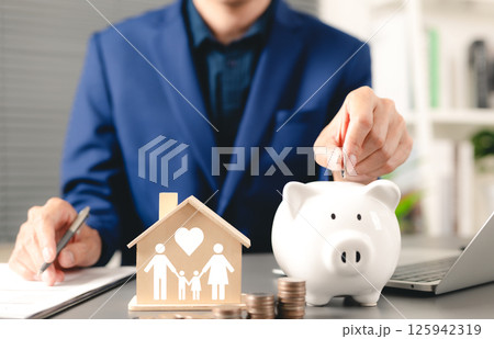Family saving money concept. Businessman deposits coin into piggy bank with wooden house model, symbolizing family and love, family financial planning, and savings. cash management, life insurance, Family saving money concept. Businessman deposits coin into piggy bank with wooden house model, symbolizing family and love, family financial planning, and savings. cash management, life insurance, 125942319
