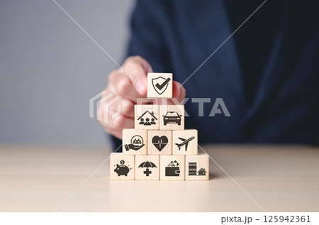 Insurance concept. Businessman arranges wooden blocks with insurance icons, financial security, health, home, and travel coverage. Top block features shield with checkmark, representing protection. Insurance concept. Businessman arranges wooden blocks with insurance icons, financial security, health, home, and travel coverage. Top block features shield with checkmark, representing protection. 125942361