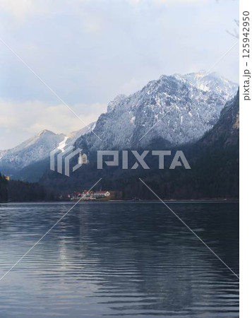 Mountainous landscape with calm lake reflecting snow-capped peaks and distant building under overcast sky Mountainous landscape with calm lake reflecting snow-capped peaks and distant building under overcast sky 125942950