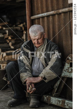 A pensioner rests on the ground, his gaze lost in thought. The elderly man wears clothes that have seen many years of work. A pensioner rests on the ground, his gaze lost in thought. The elderly man wears clothes that have seen many years of work. 125943554