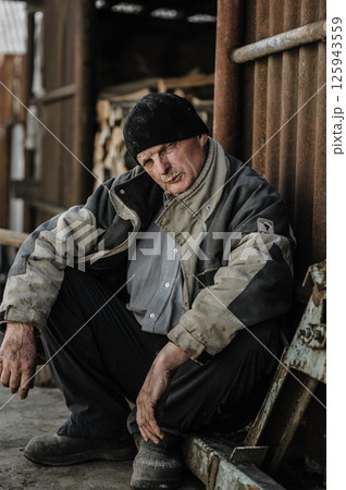 The pensioner crouches, exhausted by years of hardship. The elderly man is dressed in worn-out work clothes. The pensioner crouches, exhausted by years of hardship. The elderly man is dressed in worn-out work clothes. 125943559