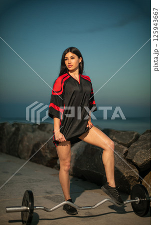 woman stands confidently one leg on barbell near shore. She wears stylish athletic attire. sun is setting casting warm glow over scene. woman stands confidently one leg on barbell near shore. She wears stylish athletic attire. sun is setting casting warm glow over scene. 125943667