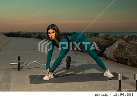 fitness enthusiast performs stretch on mat ocean showcasing determination health and exercise. Weights sit alongside workout area during sunset. fitness enthusiast performs stretch on mat ocean showcasing determination health and exercise. Weights sit alongside workout area during sunset. 125943681