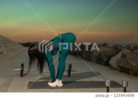 person in workout attire is executing forward bend stretch on mat located along rocky shoreline. vibrant sunset highlights sky warm hues creating serene atmosphere. person in workout attire is executing forward bend stretch on mat located along rocky shoreline. vibrant sunset highlights sky warm hues creating serene atmosphere. 125943690