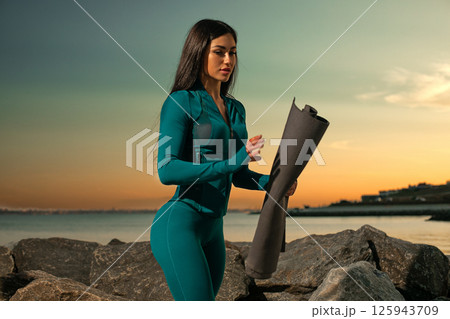fitness enthusiast rolls out yoga mat on rocky shorelines as sun sets over horizon. tranquil setting offers peaceful ambiance perfect for meditation and stretching. fitness enthusiast rolls out yoga mat on rocky shorelines as sun sets over horizon. tranquil setting offers peaceful ambiance perfect for meditation and stretching. 125943709