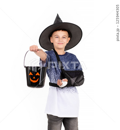 Halloween celebration concept. Portrait emotional cheerful boy in black witch hat with an injured hand, with bucket pumpkin Jack for sweet candies. Isolated white background Halloween celebration concept. Portrait emotional cheerful boy in black witch hat with an injured hand, with bucket pumpkin Jack for sweet candies. Isolated white background 125944305