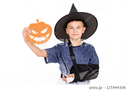 Halloween celebration concept. Portrait emotional cheerful boy in black witch hat with an injured hand, with bucket pumpkin Jack for sweet candies. Isolated white background Halloween celebration concept. Portrait emotional cheerful boy in black witch hat with an injured hand, with bucket pumpkin Jack for sweet candies. Isolated white background 125944306