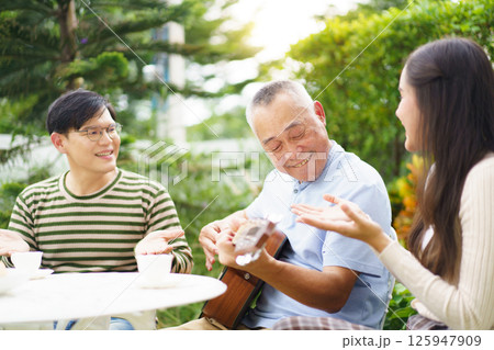 Happy Asian family spending time together, man and woman let their father playing a guitar and singing in backyard garden at home. Mental health care and wellbeing in senior adult. 125947909