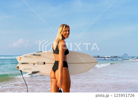 Blonde woman in black bikini holds surfboard on beach. Ocean waves in background. Ready for surfing adventure. Sunny weather, clear sky. Perfect scene for water sports, summer. Enjoys seaside. Blonde woman in black bikini holds surfboard on beach. Ocean waves in background. Ready for surfing adventure. Sunny weather, clear sky. Perfect scene for water sports, summer. Enjoys seaside. 125951726