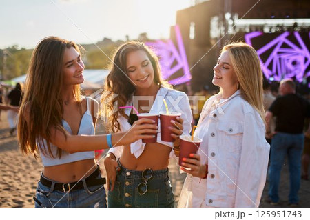 Women cheers with alcoholic beverages at sunset beach party. Smiling females holding cups, enjoying music event. Group of ladies celebrating, having fun on sandy shore near festival stage. 125951743